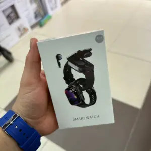 Mano sosteniendo caja blanca de smartwatch negro con audífonos inalámbricos integrados y pantalla encendida mostrando hora.